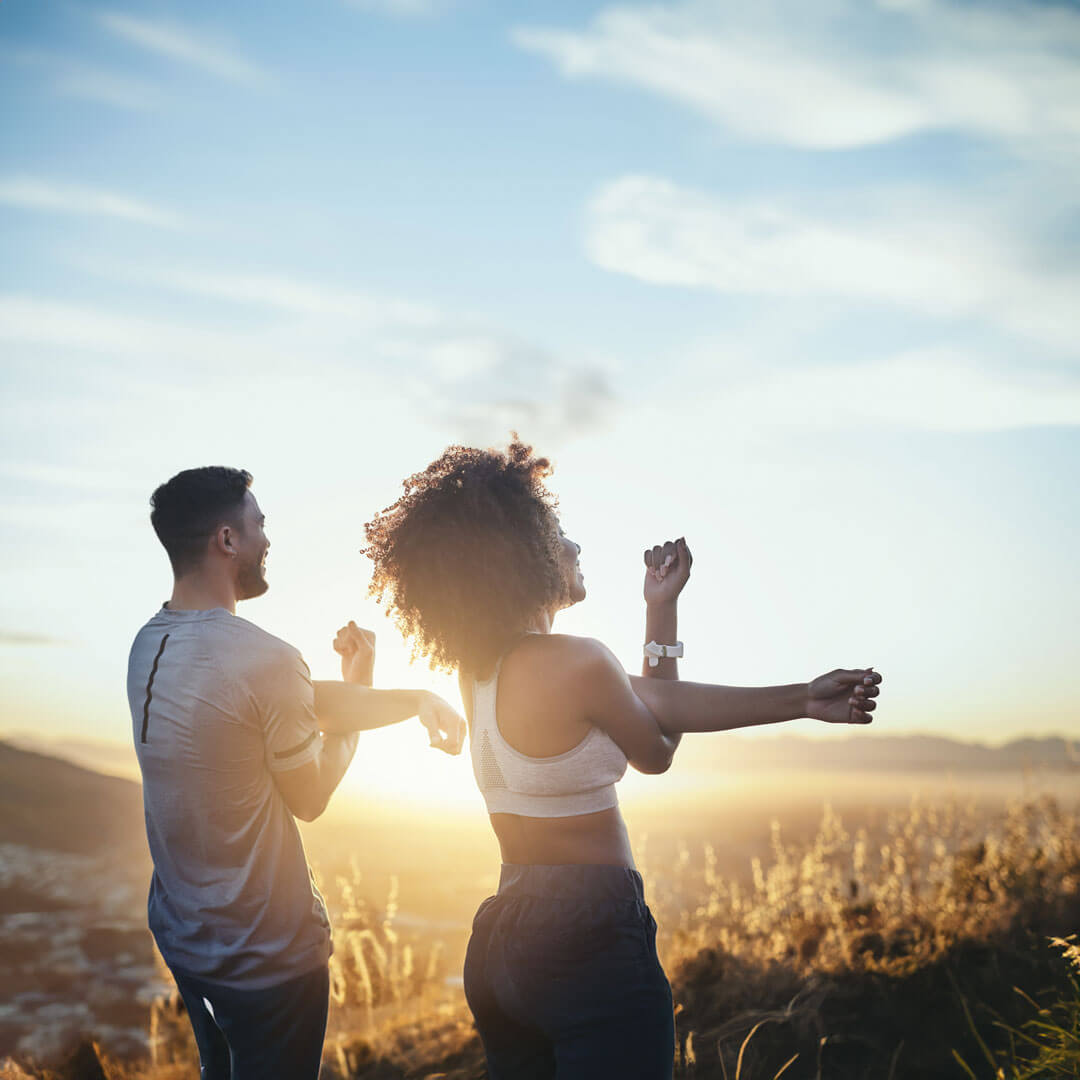 Two people stretching in workout clothes on a hillside at sunrise.