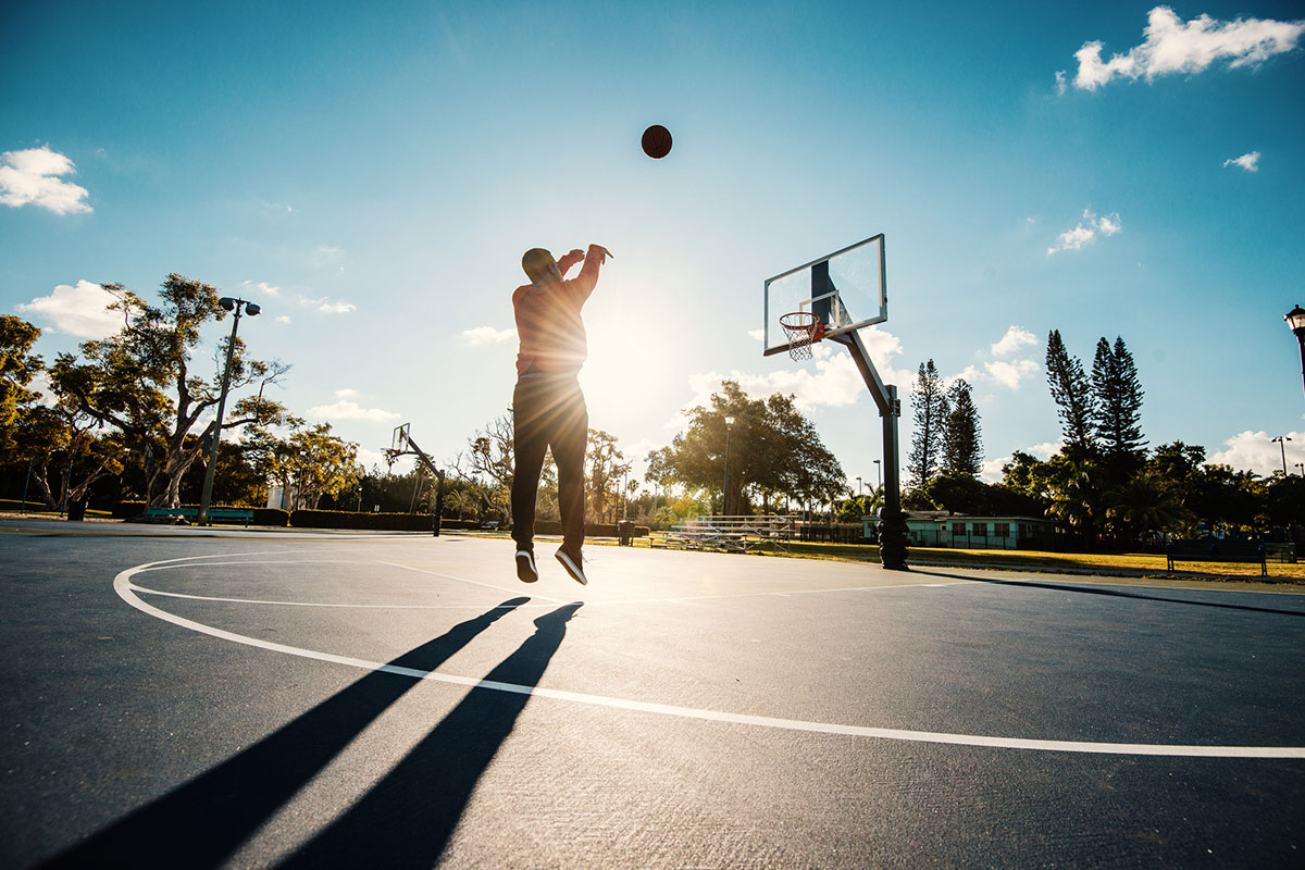 A person shoots a basketball outdoors at sunset on an empty court.