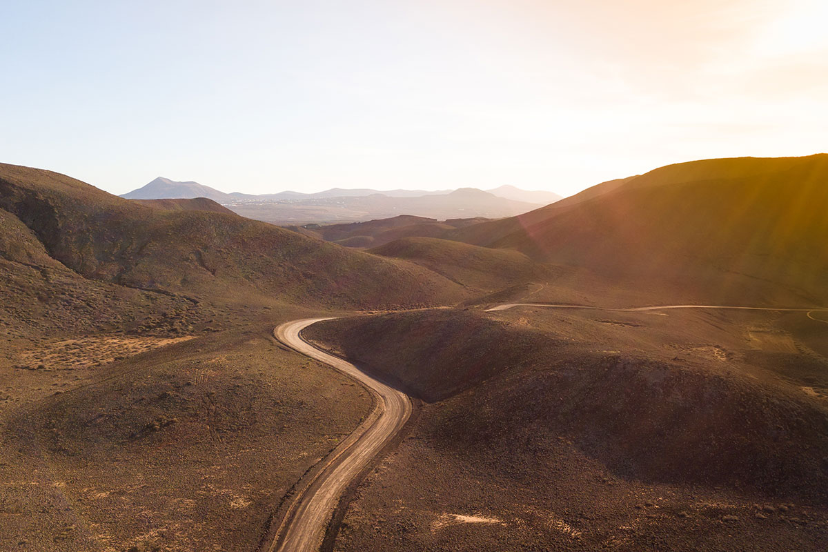 A winding dirt road snakes through sunlit desert hills under a clear sky.